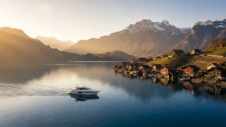 Motoryacht auf einem Schweizer Bergsee im Abendlicht, am Ufer ein Dorf mit Holzhäusern vor verschneiten Gipfeln