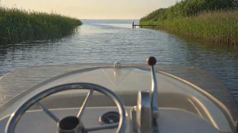 Blick vom Steuerstand eines Motorboots über den Bug auf eine schilfgesäumte Wasserstraße, die auf einen See hinausführt