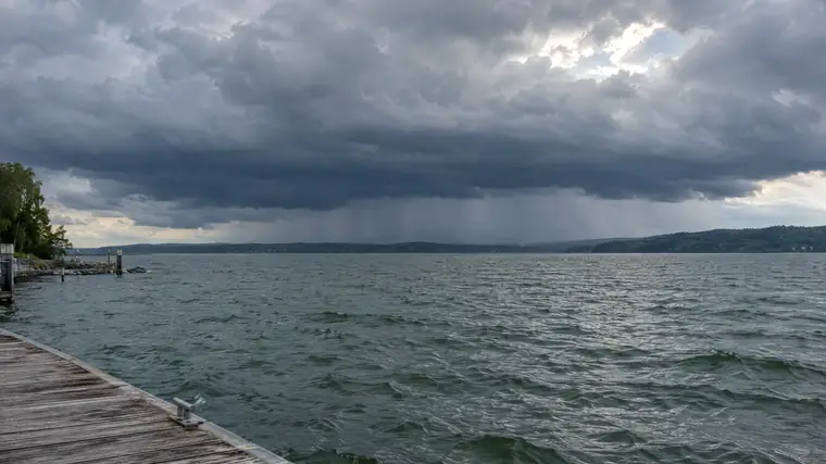 Dunkle Wolken und ein Regenschauer über einem See, im Vordergrund ein Holzsteg