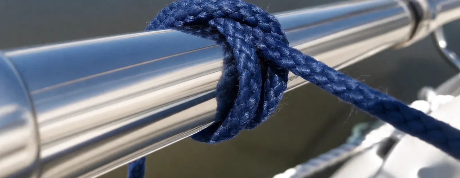 Dunkelblaue Leine mit einem Webleinstek um eine Reling aus Edelstahl, dahinter das Wasser