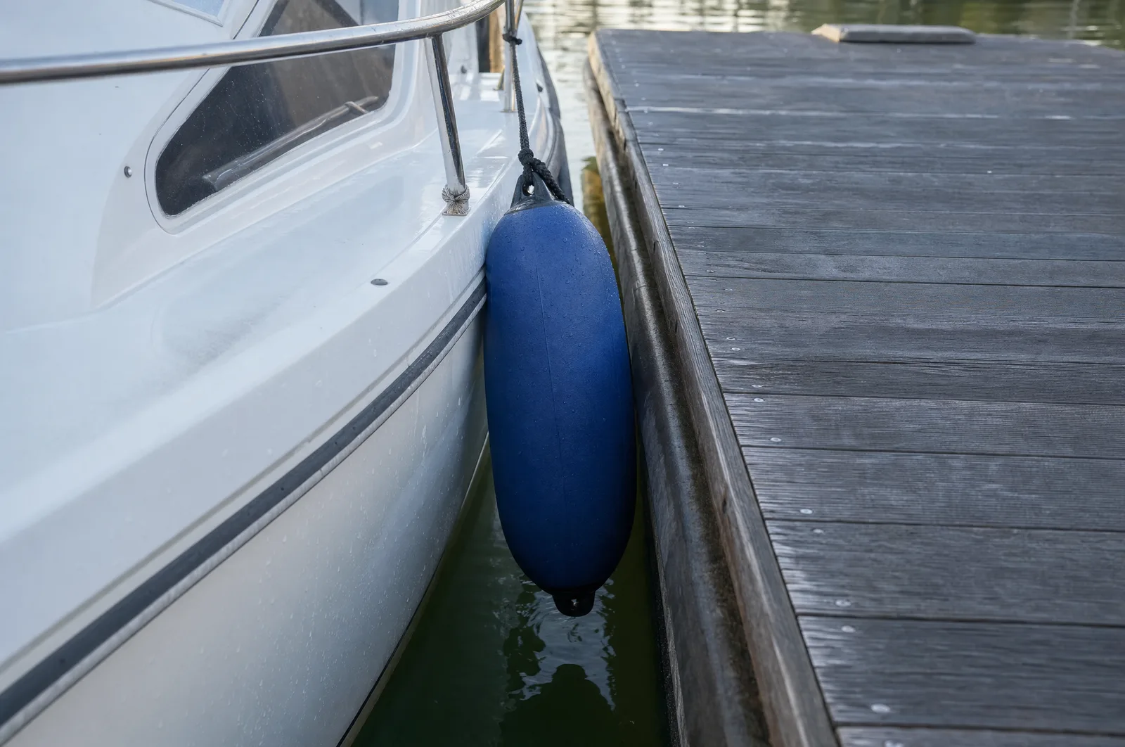 Blauer Fender hängt an einer Leine zwischen der weißen Bordwand eines Motorboots und der Kante eines grauen Holzstegs