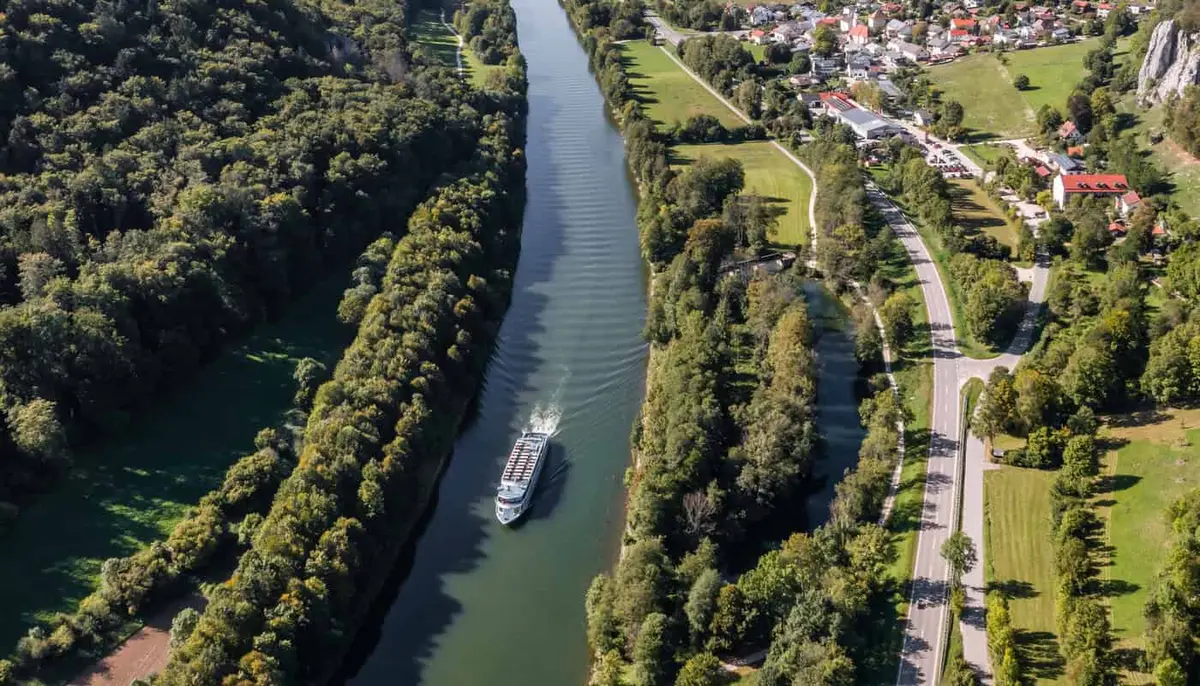 Luftbild eines Kanals zwischen bewaldeten Ufern, auf dem ein langes Fahrgastschiff fährt