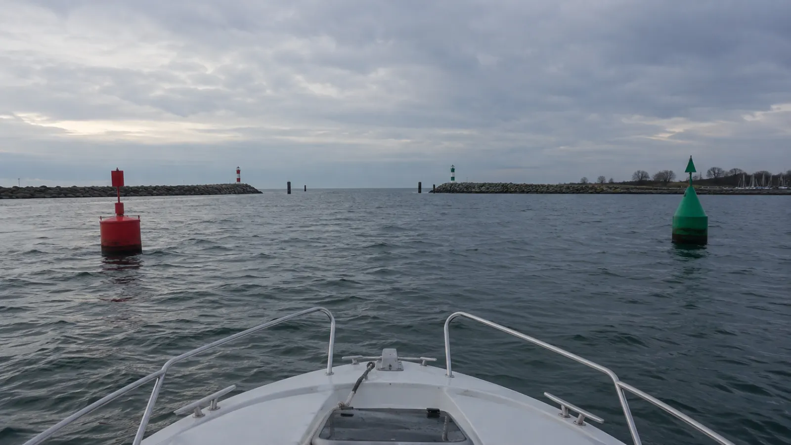 Blick über den Bug eines Motorboots beim Einlaufen in einen Ostseehafen: links eine rote Stumpftonne, rechts eine grüne Spitztonne, dahinter die Molen mit rotem und grünem Feuer