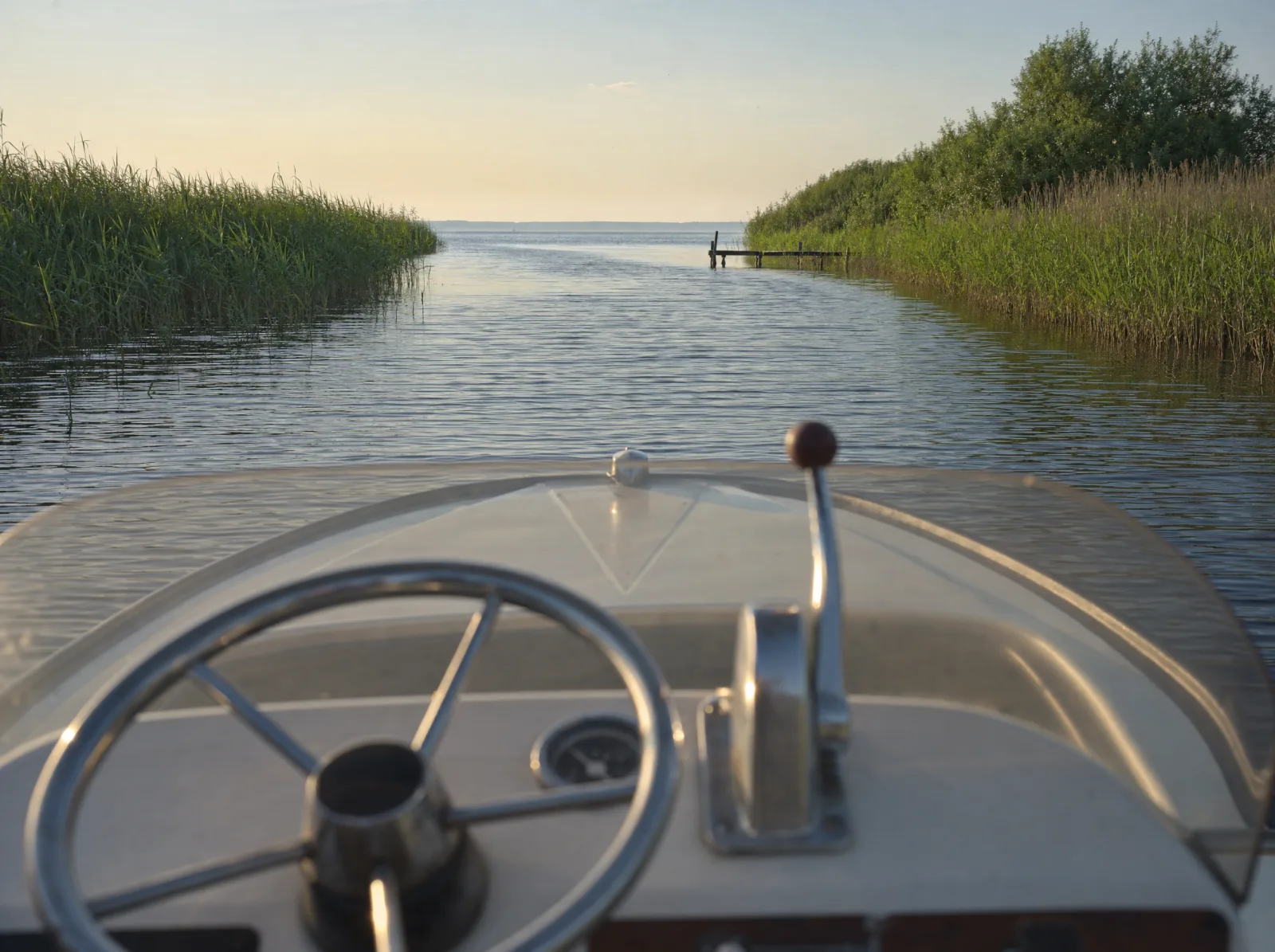 Blick vom Steuerstand eines Motorboots über den Bug auf eine schilfgesäumte Wasserstraße, die auf einen See hinausführt
