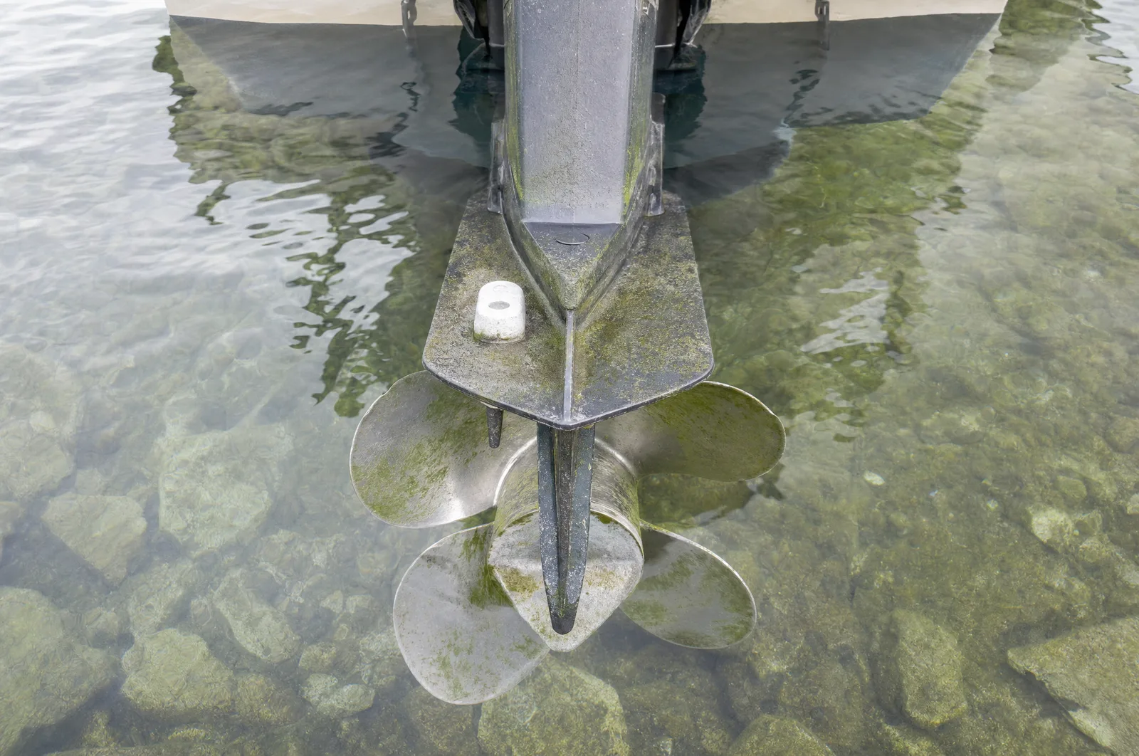Antriebseinheit eines Motorboots von hinten durch klares Wasser gesehen, Propellerblätter und Kavitationsplatte mit grünem Algenfilm, darunter Kiesgrund