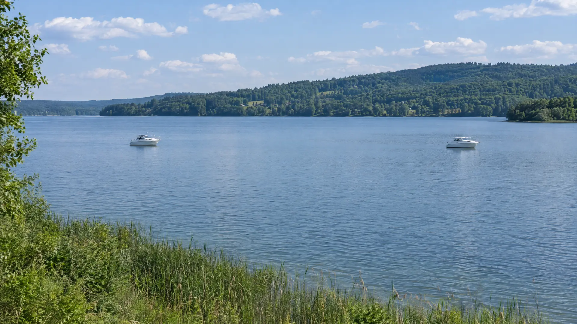 Zwei kleine weiße Motorboote liegen weit auseinander vor Anker auf einem ruhigen See, im Vordergrund Schilf am Ufer
