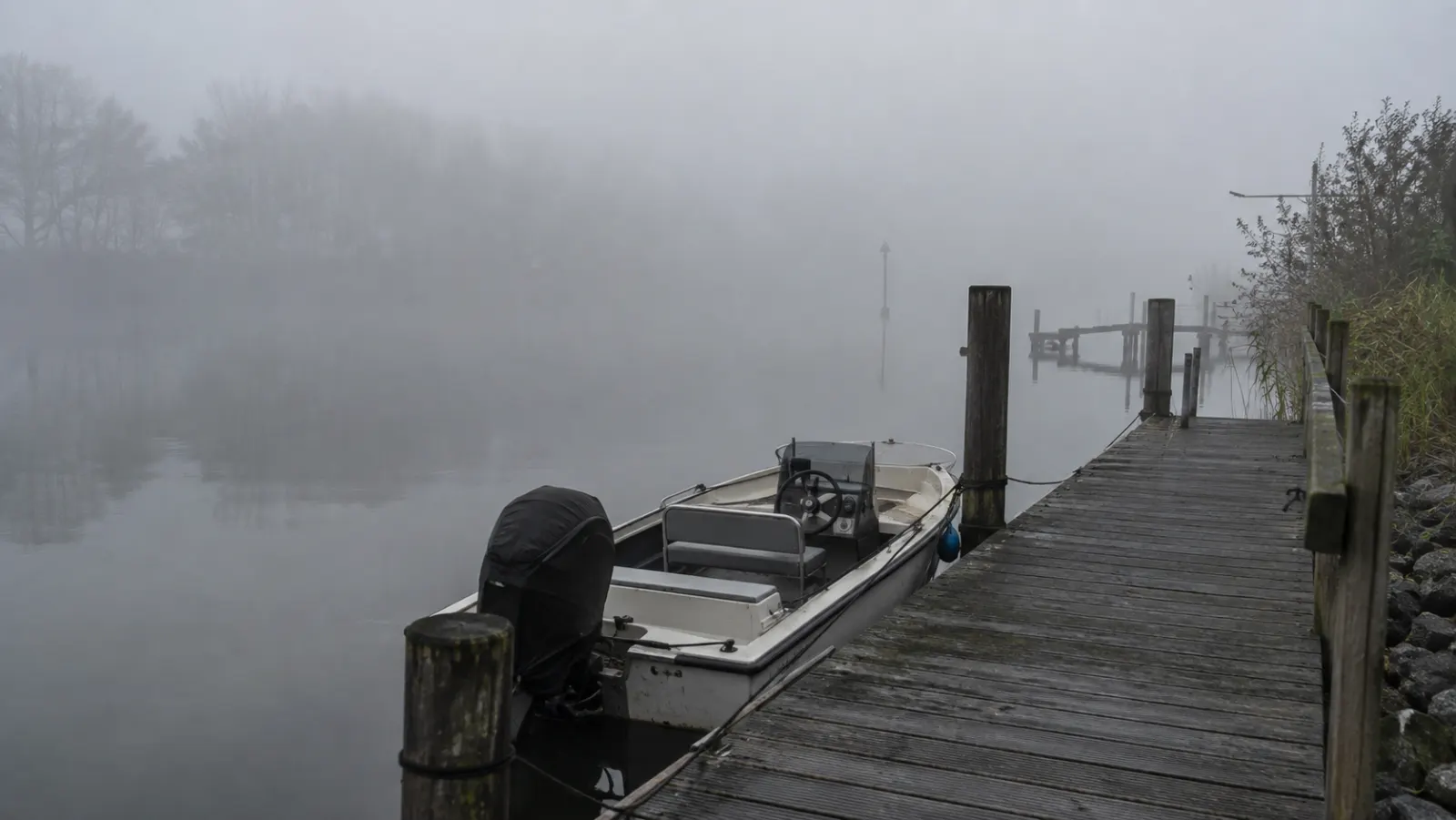 Kleines Motorboot an einem Steg am Flussufer bei dichtem Nebel, das andere Ufer ist nur schemenhaft zu sehen