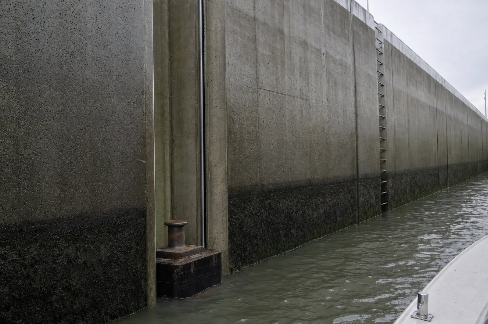 Hohe nasse Betonwand einer Schleusenkammer mit einer schmalen senkrechten Nische bis zur Oberkante. Unten in der Nische, knapp über dem Wasser, ein Schwimmpoller auf einem Stahlkasten, weiter hinten eine Steigleiter.