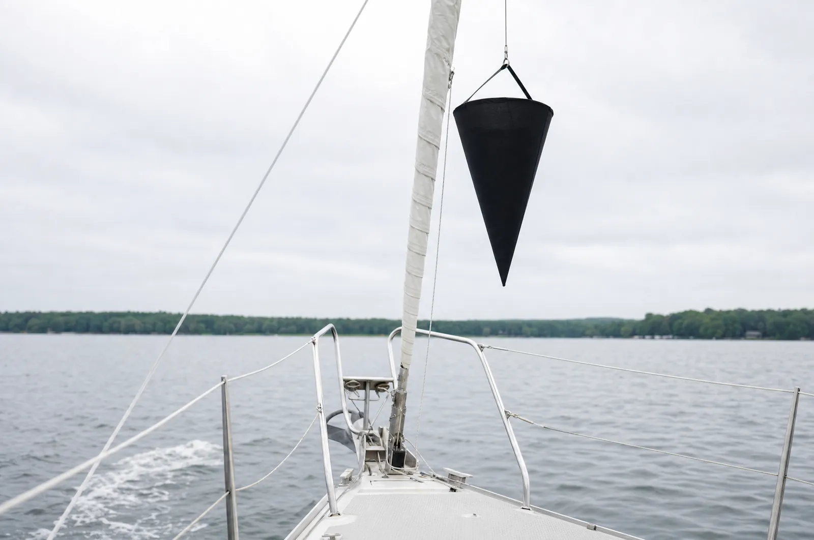 Schwarzer Signalkegel mit der Spitze nach unten hängt an einem dünnen Fall im Vorschiff einer Segelyacht neben dem eingerollten Vorsegel. Darunter Vordeck und Bugkorb, links das Kielwasser der Motorfahrt.