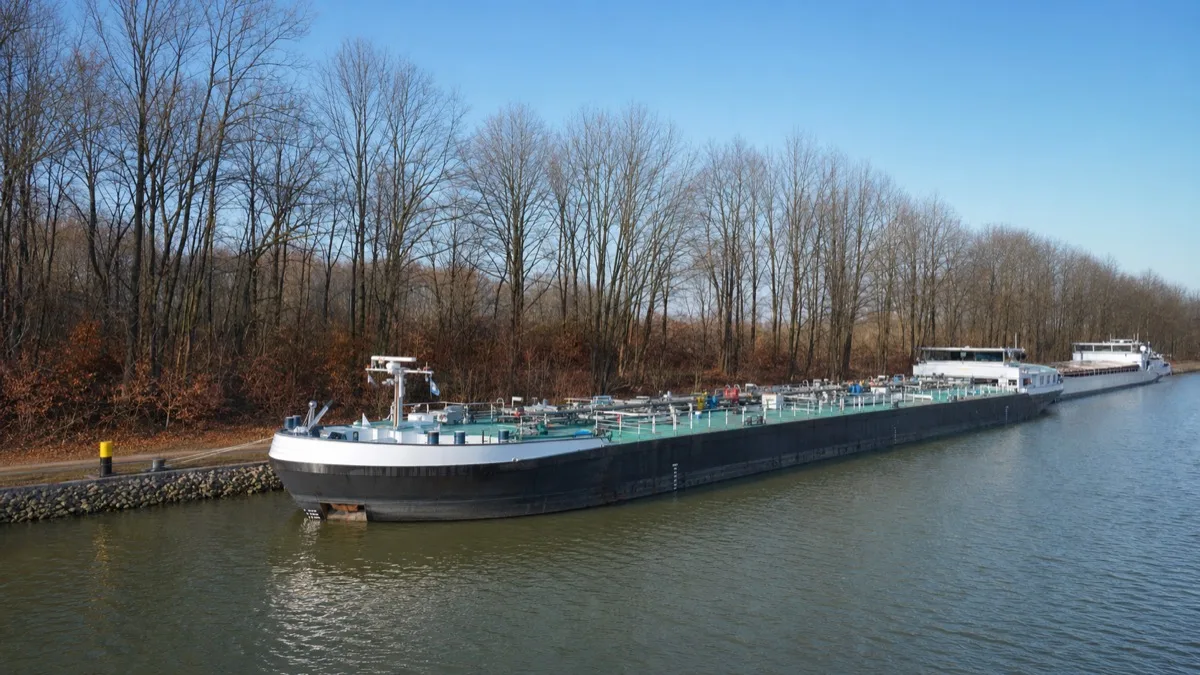 Beladenes Tankmotorschiff und zwei weitere Frachtschiffe längsseits an der befestigten Liegestelle eines breiten Schifffahrtskanals, kahle Bäume am Ufer, klarer blauer Winterhimmel