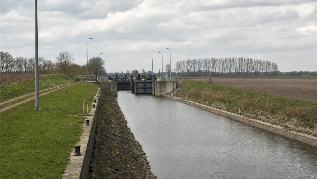 Gerader Schifffahrtskanal zwischen Feldern, links ein Grasdamm mit Pollern und Laternen, in der Mitte eine Schleuse mit stählernen Toren unter bedecktem Himmel