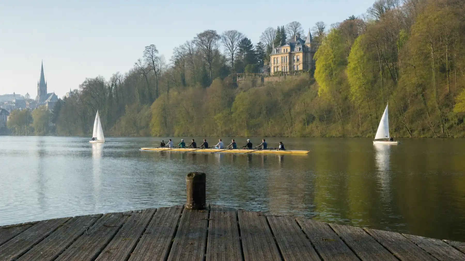Schmaler aufgestauter Fluss am Morgen, das bewaldete Gegenufer nur wenige hundert Meter entfernt: auf dem Wasser ein Ruderachter und zwei kleine Segeljollen, am Hang eine große Villa, links die Türme der Stadt