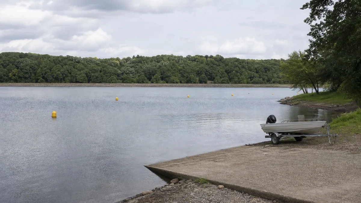 Slipanlage an einem aufgestauten Fluss: ein kleines Motorboot auf dem Trailer am Ufer, auf dem Wasser gelbe Bojen, gegenüber bewaldetes Ufer