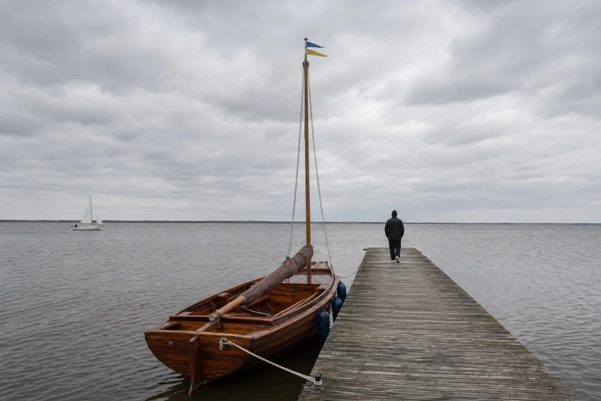 Offenes Holzboot mit breitem, flachem Rumpf und stehendem Holzmast längsseits an einem Holzsteg, dahinter ein weiter Binnensee unter grauem Himmel mit einer Segelyacht in der Ferne
