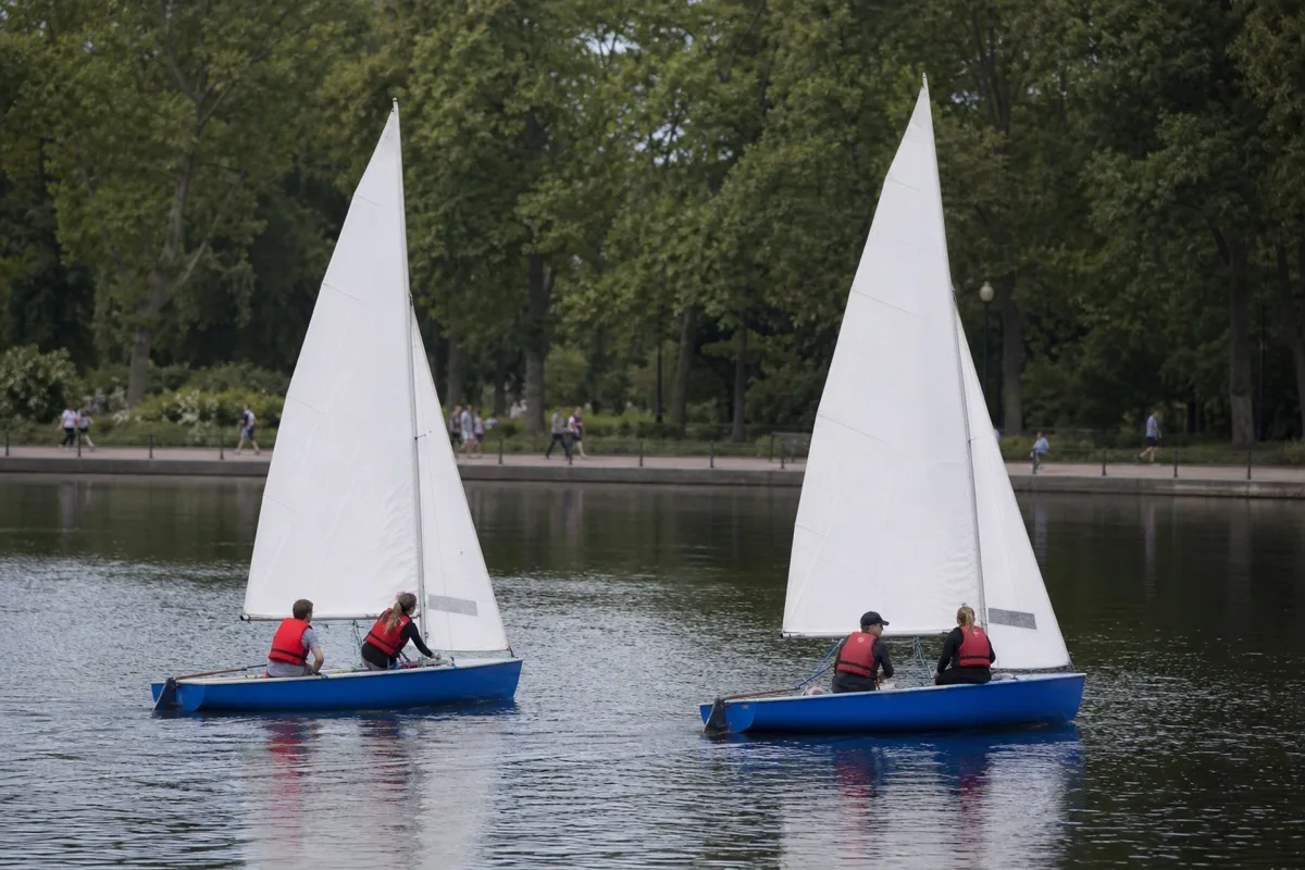 Zwei blaue Segeljollen mit weißen Segeln auf einem Stadtsee, die Crews in roten Rettungswesten, dahinter eine baumbestandene Uferpromenade