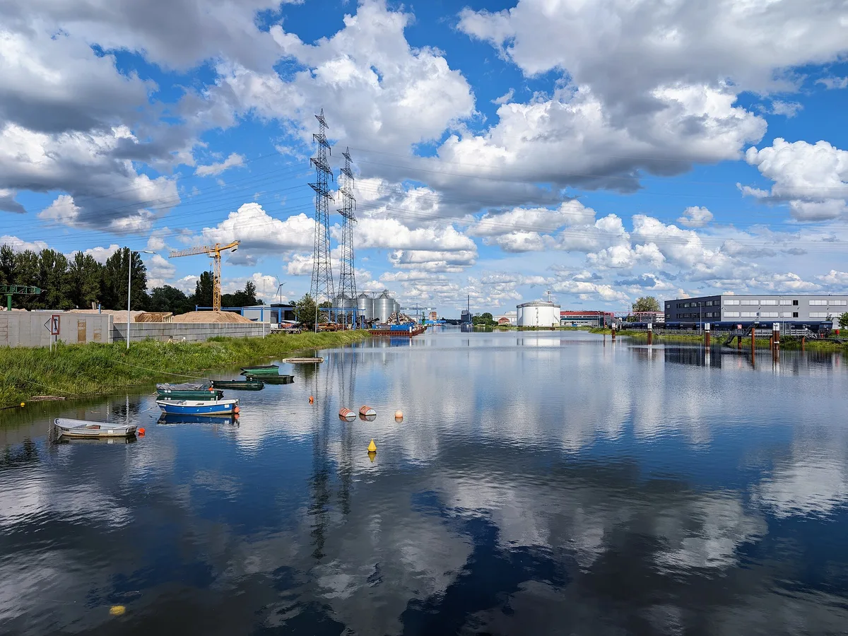 Hafenbecken der Rheinhäfen Karlsruhe mit spiegelglattem Wasser, links kleine Boote am Ufer, dahinter Silos, Tanks und Strommasten unter Quellwolken