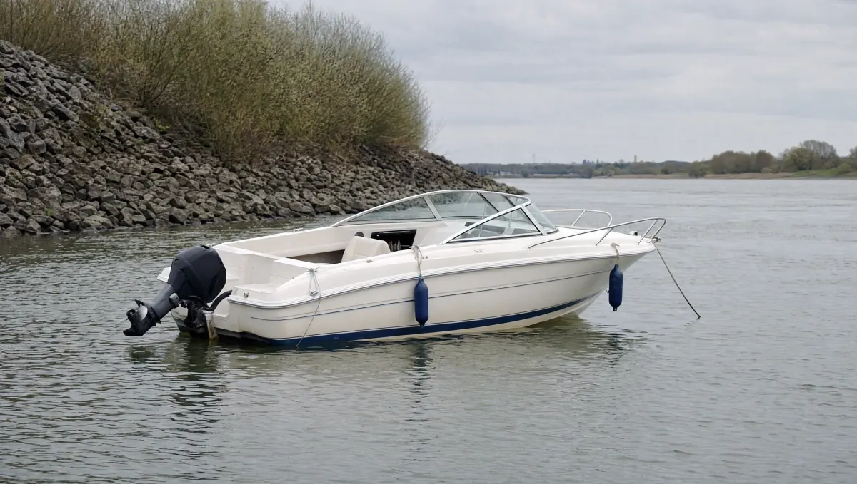 Weißes Sportmotorboot mit blauen Fendern vor Anker auf einem breiten Fluss, dahinter eine grobe Steinschüttung mit Weidengebüsch