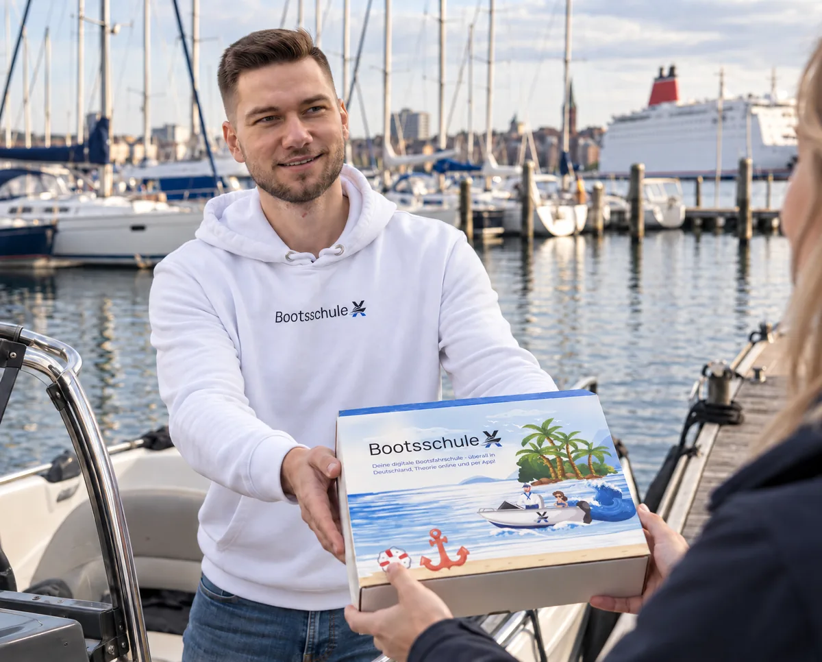 Mann im BootsschuleX-Pullover reicht die Willkommensbox von einem Motorboot an eine Frau auf dem Steg, dahinter ein großer Yachthafen und eine Fähre