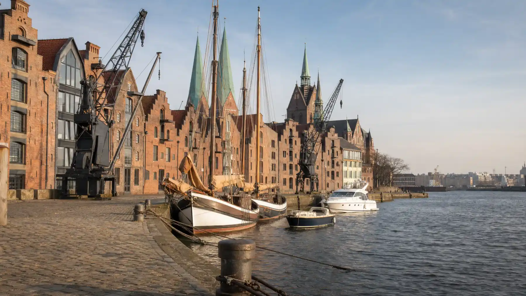Rote Backsteinspeicher mit Staffelgiebeln und zwei schwarze Hafenkräne an der Kaimauer, davor zwei hölzerne Traditionssegler, eine weiße Motoryacht und ein kleines offenes Boot