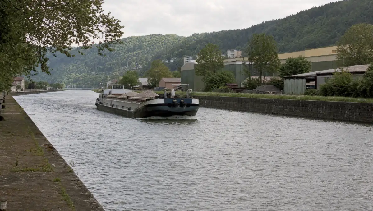 Beladenes Frachtschiff in Fahrt auf einem zwischen Ufermauern gefassten Fluss, bewaldete Hänge und Werkhallen am Ufer