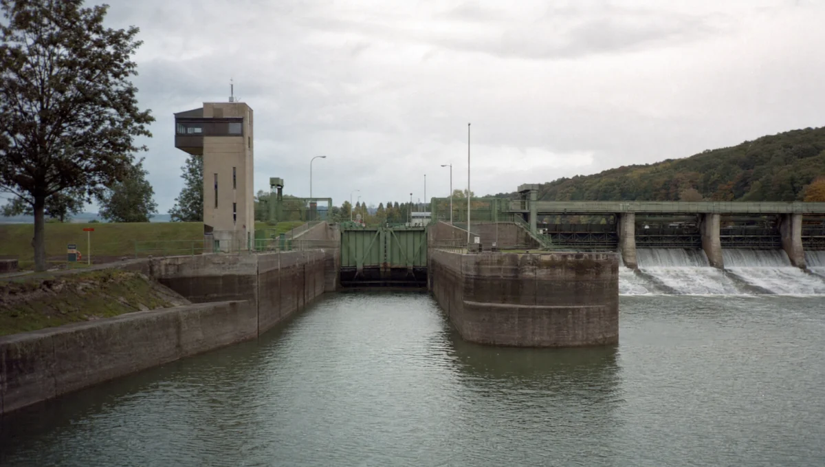 Flussschleuse mit langer Betonkammer und grün gestrichenen Stahltoren, daneben ein Steuerhaus und ein mehrfeldriges Wehr